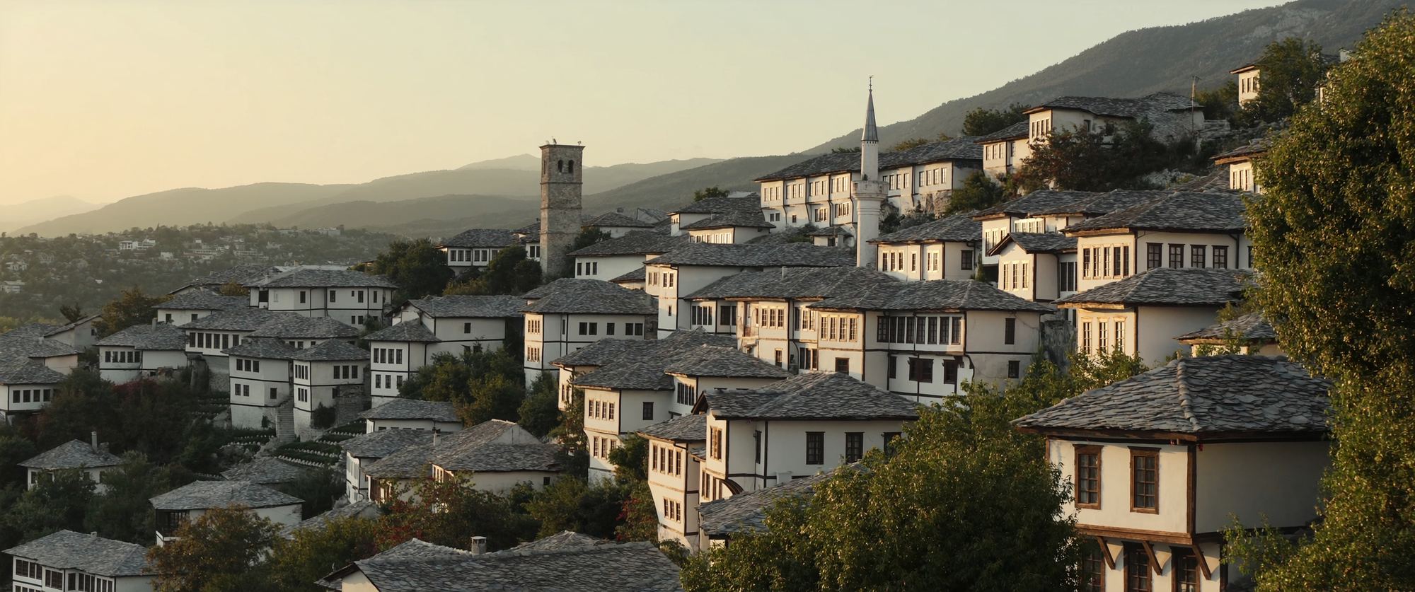 An old hill town of white stone houses with grey slate roofs climbing a slope, with a minaret and a stone tower, at golden hour